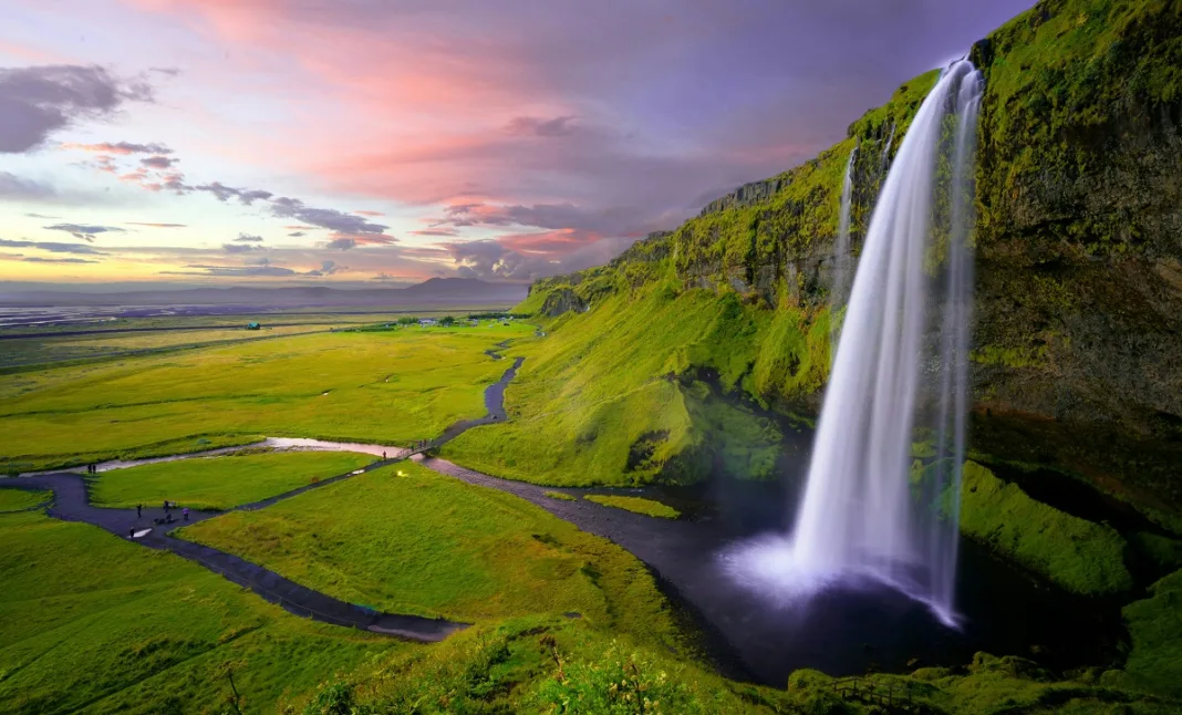 Scenic view of a tall waterfall cascading down a moss-covered cliff into a serene pool at sunset, creatively representing “waterfalls in Dallas” with vibrant green landscape and colorful sky.