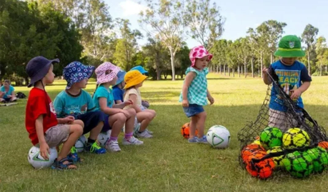 Group of young children wearing colorful hats sitting on soccer balls in a park while two toddlers play with more balls from a net bag during a soccer activity session.